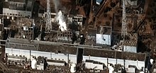 Photographie couleur d'une centrale nucléaire en ruine, vue de dessus, avec de la fumée qui se dégage des ruines.