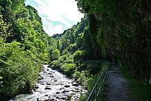 Sentier le long d'une falaise qui donne sur une rivière, le tout entouré de verdure.