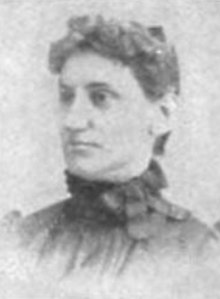 A white woman with curly dark hair in an updo, wearing a high-collared dark dress or blouse.