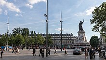 Frontale Farbfotografie eines Platzes mit einer Straße im Vordergrund, auf der rechts zwei Autos fahren. Rechts ist eine große, dunkle Statue mit Toga und Zweig in der rechten Hand. Sie steht auf einem hohen, weißen Rundpodest, das in der Mitte von weißen Figuren umschlossen wird und unten mit Graffiti beschmiert ist. Viele Menschen sind unterwegs und links hinten sind Bäume und Geschäfte.