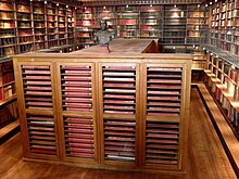 Grande salle aux murs couverts d'étagères de livres, parcourues par une galerie. Au centre, un grand meuble en bois contenant de grands registres
