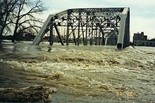 Dirty flood water rushing through the roadway of a bridge.