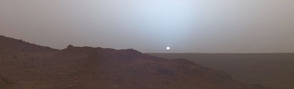 Zdjęcie panoramiczne. Na pierwszym planie skaliste wzniesienie, za nim równina. Na horyzoncie okrągła biała tarcza Słońca z niebieską poświatą.