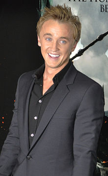 Frontale Farbfotografie von einem jungen Mann, der in die Kamera strahlt. Seine blonden Haare sind nach oben gestylt und er trägt einen schwarzen Anzug. Im Hintergrund steht ein Filmplakat.