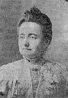 A middle-aged white woman, dark hair in an updo, wearing a white lacy blouse with a high collar