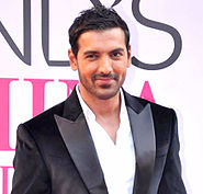 A smiling John Abraham, in a white shirt and black coat