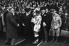 Homme à barbe blanche portant un haut-de-forme remettant une coupe à un footballeur, avec une foule autour et derrière lui