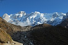 A shining white snow-clad range, framed against a turquoise sky. In the middle ground, a ridge descends from the right to form a saddle in the centre of the photograph, partly in shadow. In the near foreground, a loop of a road is seen.