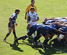 Un joueur en train de conserver le ballon dans les pieds en troisième ligne d'une mêlée fermée. Les deux demi de mêlée suivent la progression de celle-ci, l'arbitre est attentif.