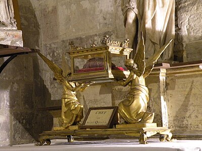 Deux anges féminins ailés et dorés tiennent une boîte présentant des ouvertures vitrées sur les côtés et contenant un morceau de tissu posé sur un coussin.