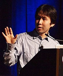 A Japanese man gestures in front of a podium.