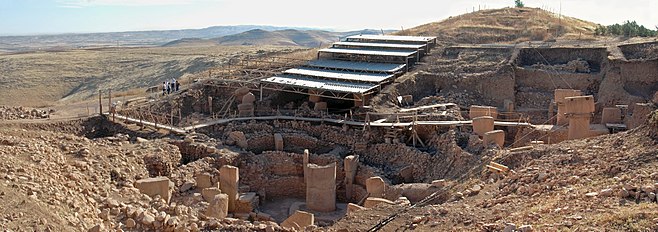 Vue panoramique du site archéologique sous un ciel bleu. À l'arrière plan, on voit l'abri de fouilles au soleil.