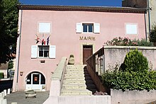 Façade rose à un étage et trois fenêtres, escalier montant la porte centrale. En bas à gauche, à côté de l’escalier, ouverture en arche dans un étage en-dessous.