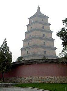 Fotografie vysoké šedobílé pagody,za zdí v popředí je vidět šest postupně se zužujících pater a věžičkou na nejvyšším patru; na každé straně každého patra je vysoké klenuté okno