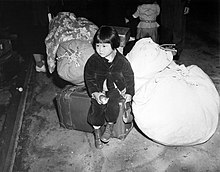 A small Asian girl is seated on luggage and large bundles, holding a partially eaten apple.