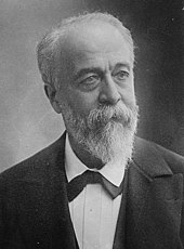 Photo en noir et blanc d’un homme ayant les cheveux blancs, une barbe taillée en pointe et habillé en costume trois pièces, avec un nœud papillon