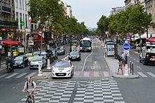 Frontale Farbfotografie einer mehrspurigen Allee mit zwei Busfahrstreifen in der Mitte, auf denen ein Bus und zwei Taxis fahren. Auf beiden Straßeninseln sind Haltestellen. Links fahren viele Autos und unten links ist ein Fahrradfahrer. An beiden Seiten sind Häuserfassaden mit Geschäften.