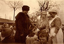 à gauche Henriette vêtue d'un manteau et d'une toque en fourrure souriante regarde un enfant qui reçoit un présent de la reine Élisabeth coiffée d'un bandeau aux côtés d'une infirmière souriante de profil