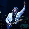 A man wearing a black tie and white dress shirt with an electric guitar hanging from a strap around his neck. His left arm is raised, with his index finger pointed upwards. In the background is a drum set.