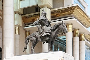 Statue of an armoured man, in front of a large pillared building.