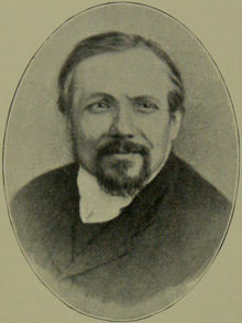 A vintage oval portrait of a man with short hair, a moustache, and a goatee, wearing a high-collared shirt and a dark coat, smiling slightly at the camera.