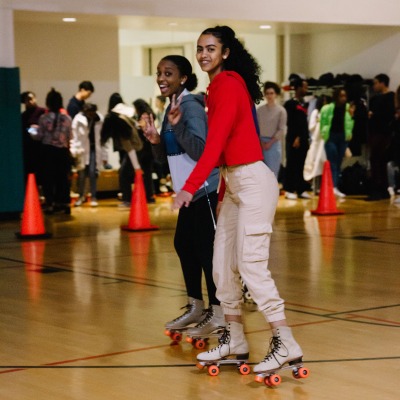 Two students roller skate together in a Rec gym with smiles on their faces.