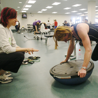 A woman balances her arms on a balance board as her trainer watches her form.