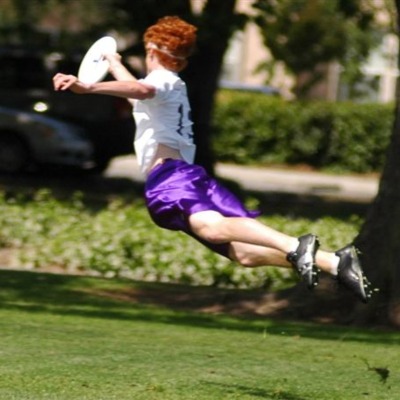 An ultimate disc play leaps through the air to catch a disc. The player wears purple shorts and has bright red hair.