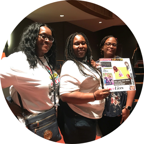 Three Black feminine presenting people, one of whom is holding up a poster.