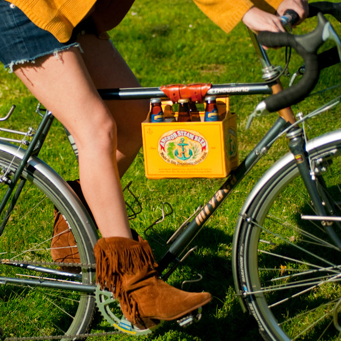 A woman bicycling on a bright summer day over a green lawn wearing suede fringe cowgirl boots and with a 6-pack of Anchor Steam beer connected to the top tube of her bicycle with a honey leather 6-pack frame cinch