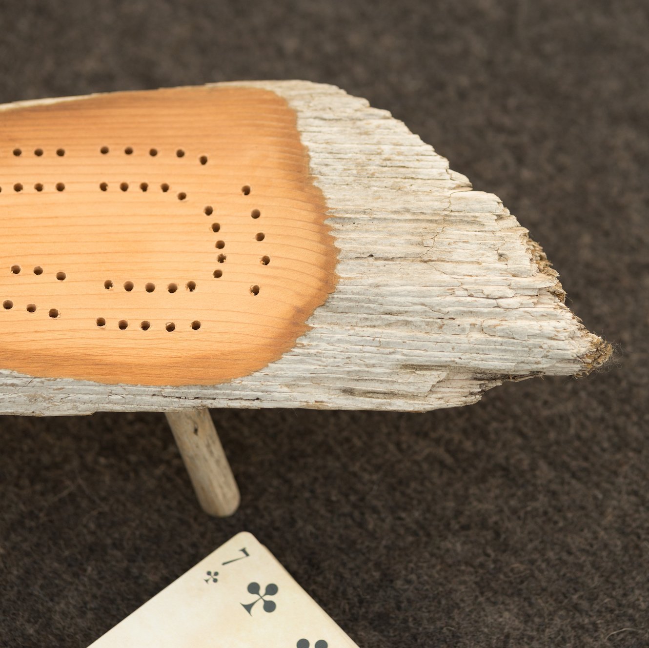 Close-up of the end of a cribbage board made from driftwood on a soft gray wool tablecloth with a vintage playing card