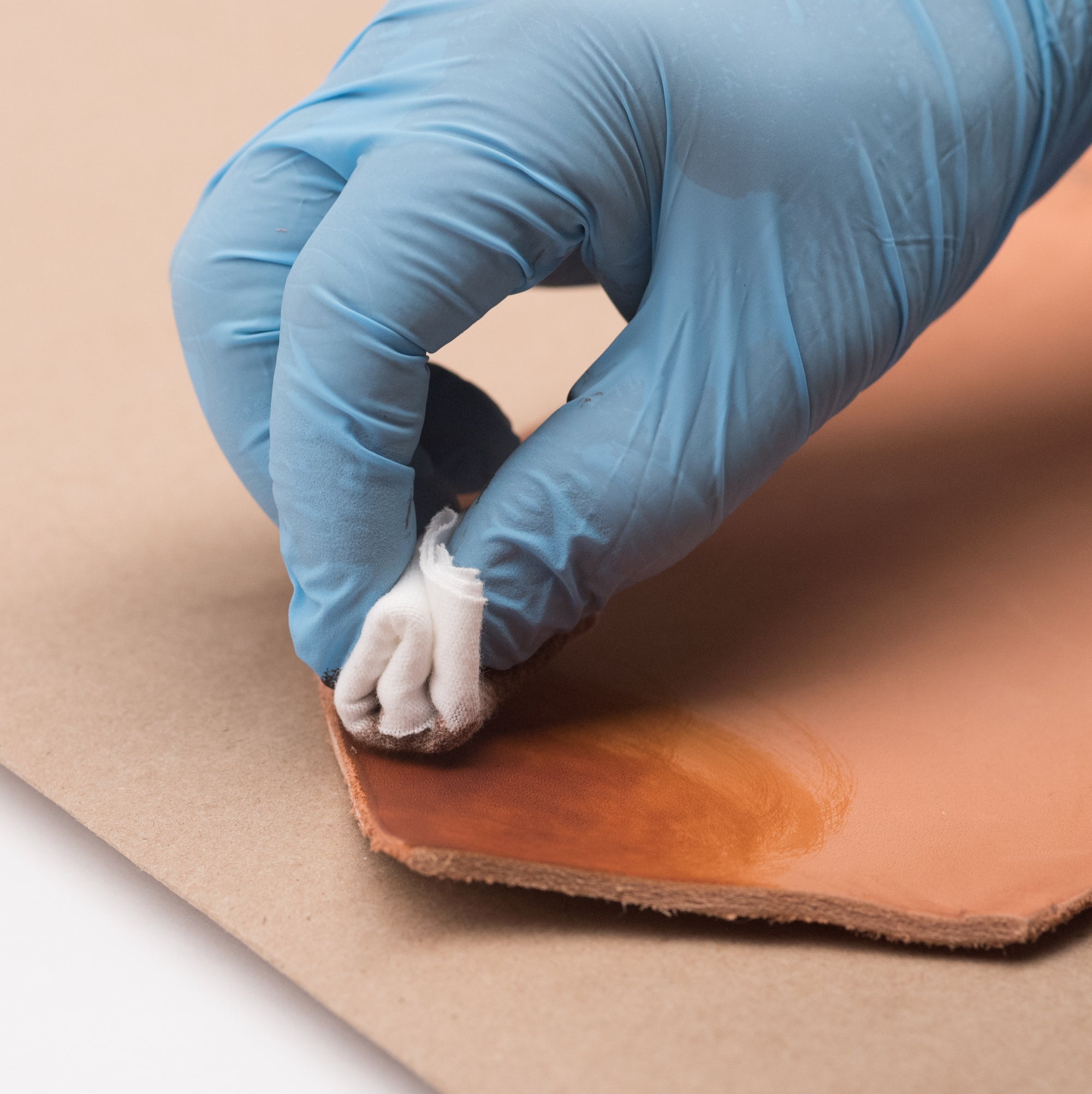 A blue-gloved hand using a small folded rag to rub dye or stain into natural leather in a circular motion on top of a kraft paper surface protector in bright natural light