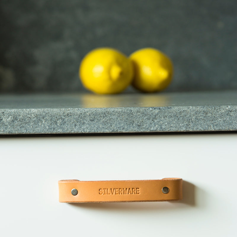 A white kitchen drawer under a gray countertop that has a natural tan colored leather handle with nickel hardware and an engraved label that says 'Silverware'