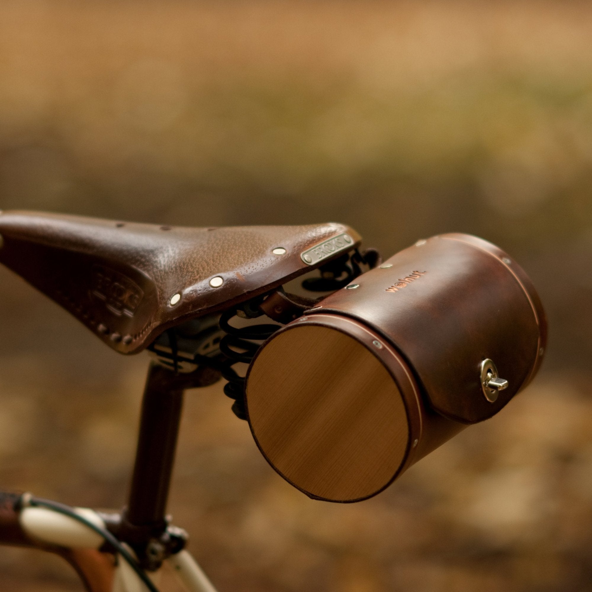 A dark brown leather round, barrel-shaped saddle bag with cedar wood sides mounted to the back of a brown leather Brooks saddle on a cream-and-brown painted bicycle