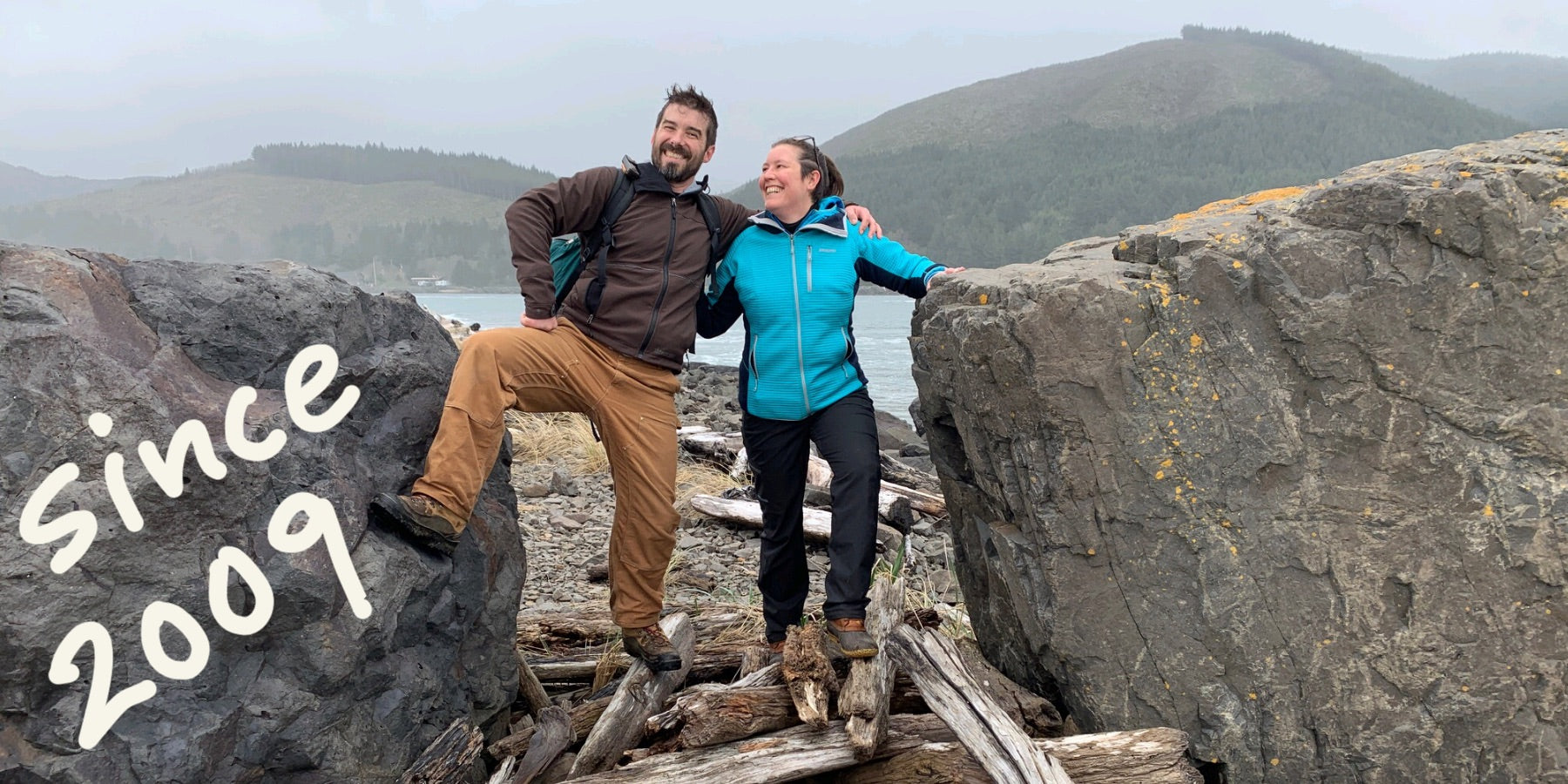 Valerie and Geoffrey Franklin of Walnut Studiolo wearing rain gear on a stormy day on the Oregon Coast beach near the jetty. They are standing on a pile of driftwood between two massive boulders that are as high as their shoulders. The mountains are misty in the background. And the words, Since 2009, are handwritten in white letting on top of the photo