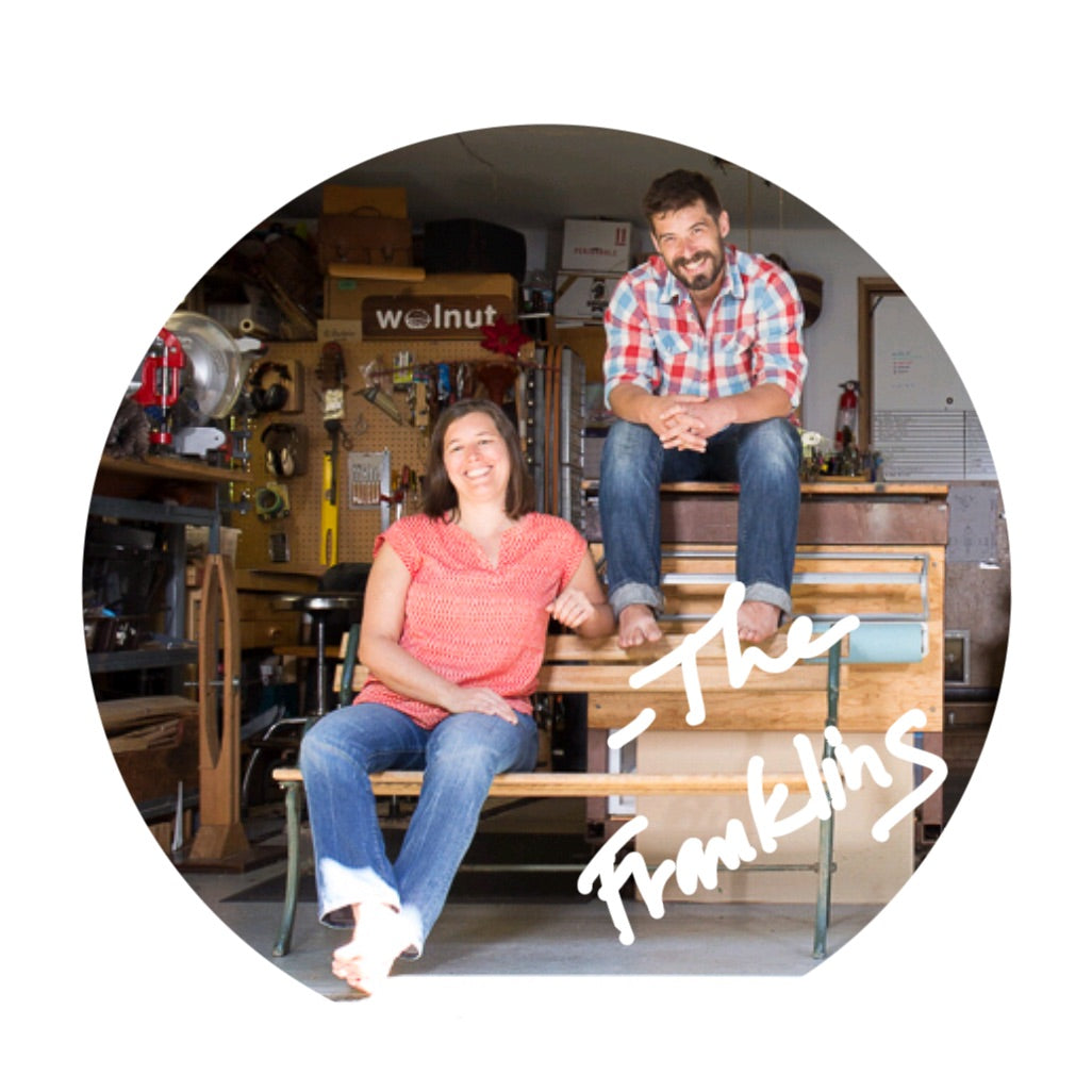A man and a woman sitting casually on a bench in a leather workshop based in a home garage in Oregon, smiling a the camera