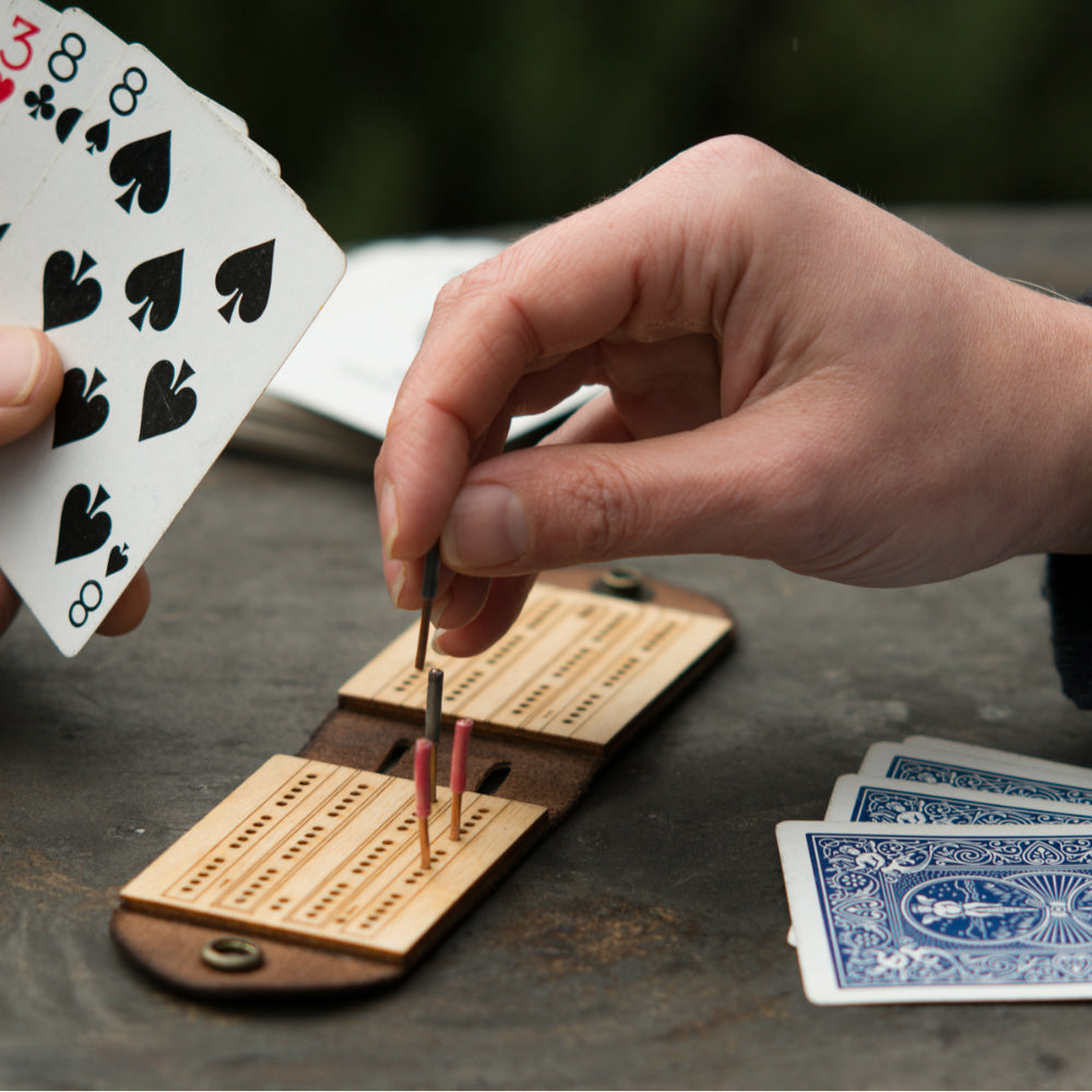 Two people playing a game of travel cribbage on a wooden picnic table with a small leather and birch cribbage board