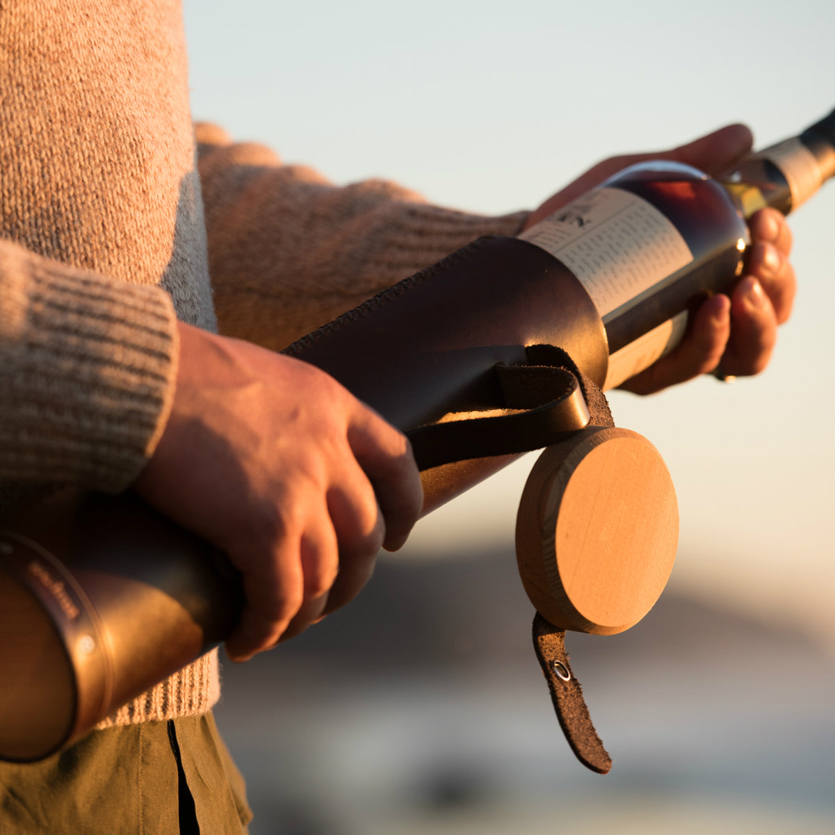 A man in a tweed sweater is pulling a bottle of Oban scotch whisky out of a handcrafted leather tube with an attached wood lid at sunset on the beach