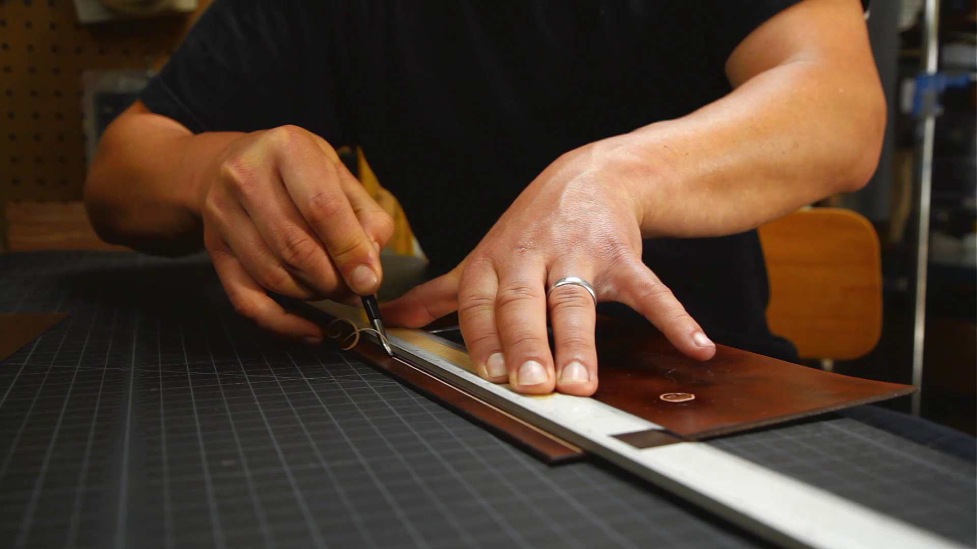 Geoff Franklin from Walnut Studiolo cutting away the surface of a dark brown dyed piece of leather to reveal the natural leather underneath with a special leather handworking tool on a black gridded cutting mat. Photo is a still from the video about handmade by Walnut from Cineastas PDX