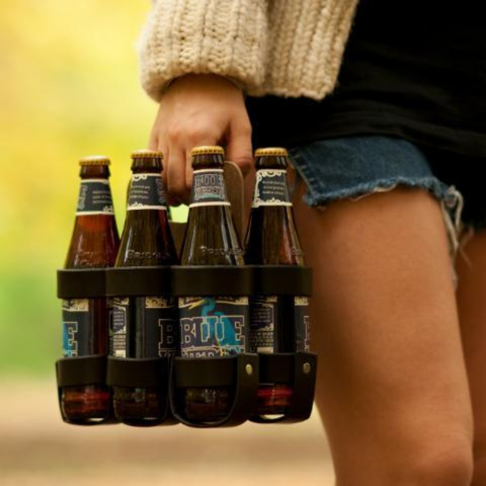 A woman wearing fashionably ripped jean shorts is carrying a leather and wood six-pack of beer on a fall day in the park