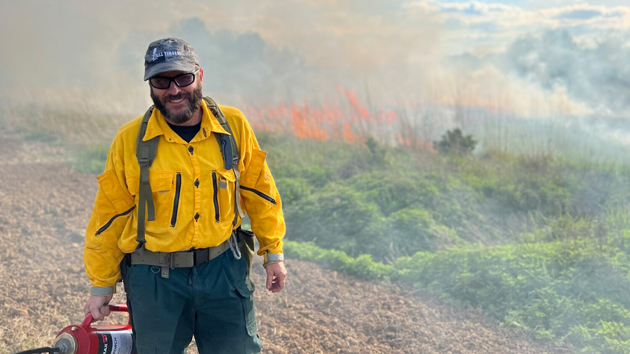 A man stands in a forested area surrounded by fire