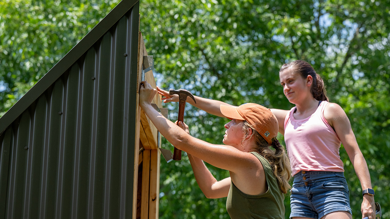 Two young women use a hammer and nails to build a structure out of wood