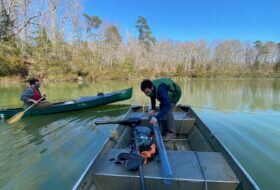 Professor Nick Balascio stands in a small boat, examining a plastic tube of sediment.