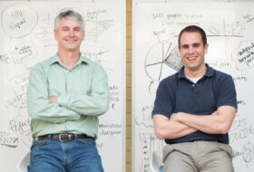 Mike Tierney and Brad Parks sitting in front of large whiteboards with detailed flow charts and graphs illustrated