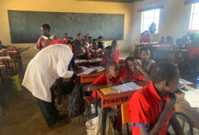 A windowed classroom with a teacher helping students dressed in school uniforms at their desks