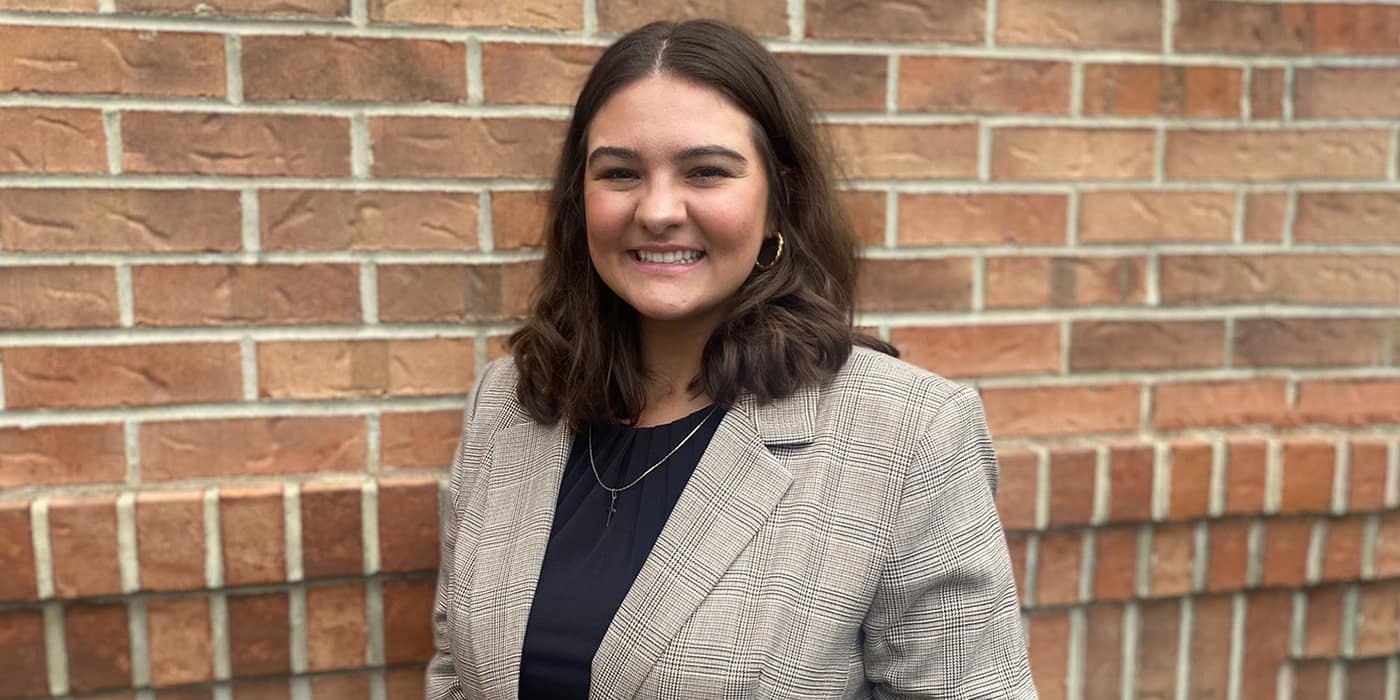 Hannah, wearing a grey plaid blazer, poses in front of a brick wall.