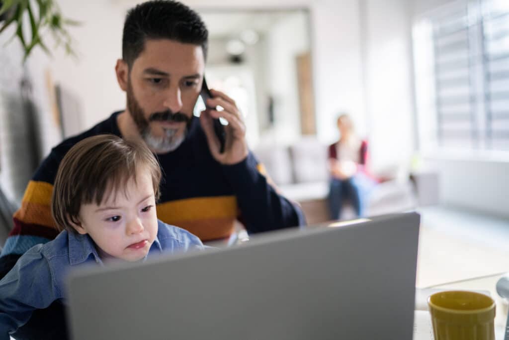 Father with beard on mobile phone looks over the shoulder of his neurodiverse son who is using a laptop.