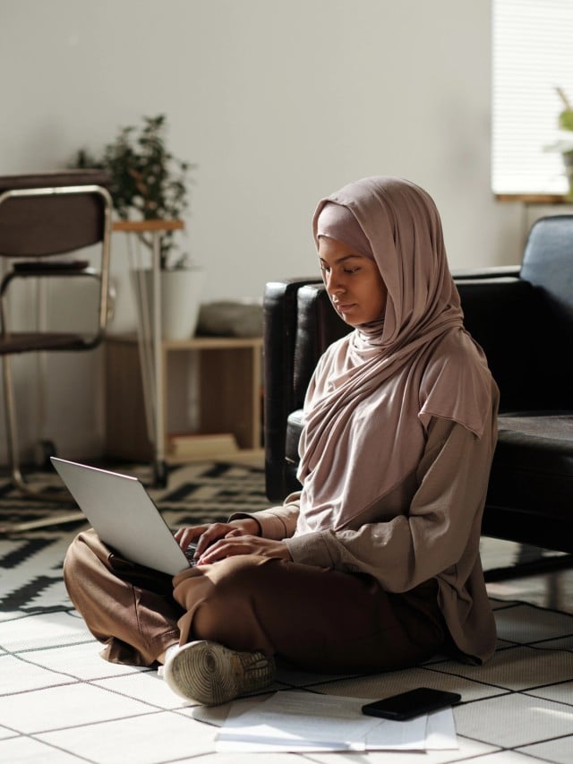 A young woman wearing a headscarf, seated on the floor and working on a laptop