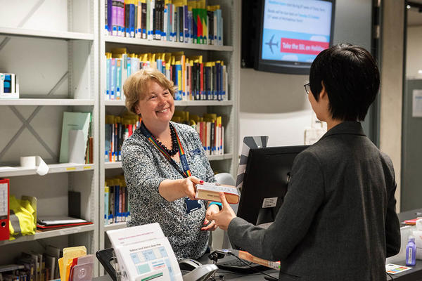 A woman in a grey blouse behind a library desk passes a book to a woman in a dark blazer