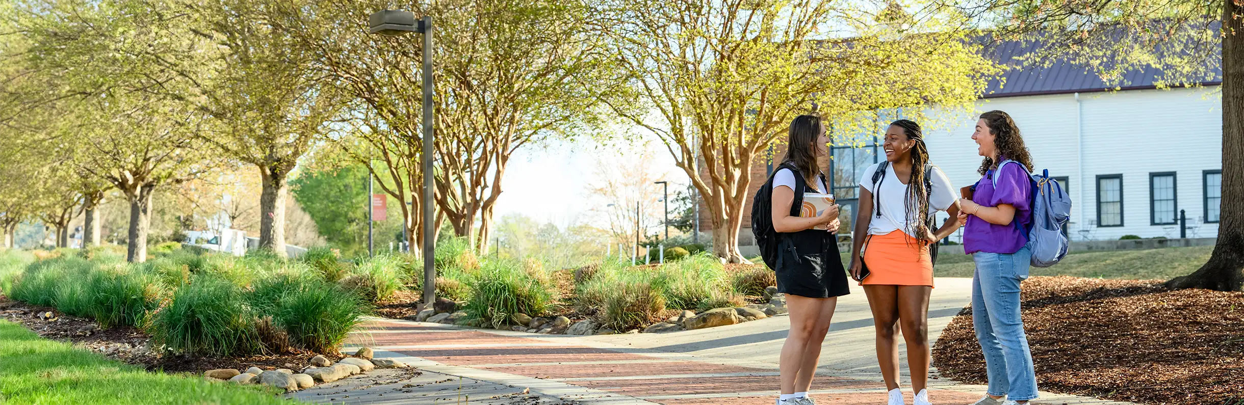 Three female students with backpacks stand on a walkway in the middle of campus in front of a painted white and brick building and green leafy trees.